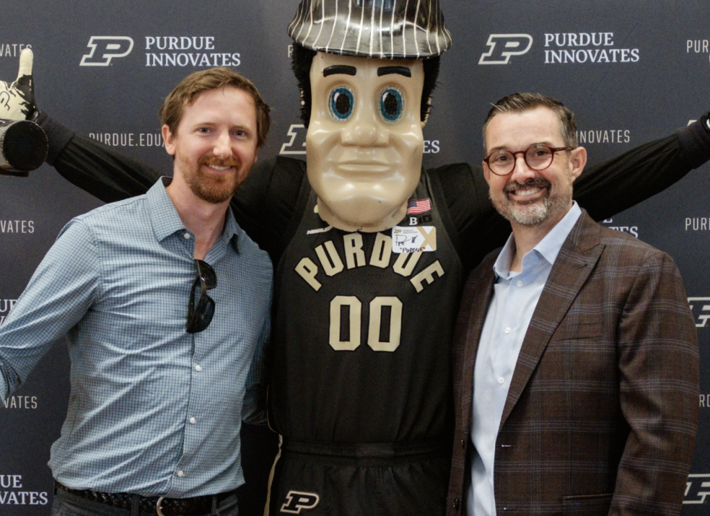 Two men and the Purdue mascot between them pose in front of a Purdue Innovates sign.